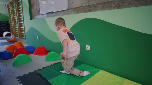 A young boy actively participates in various kids fitness activities within a colorful indoor gym. He joyfully performs a somersault, balances on soft pods, and crawls through a vibrant play tunnel.