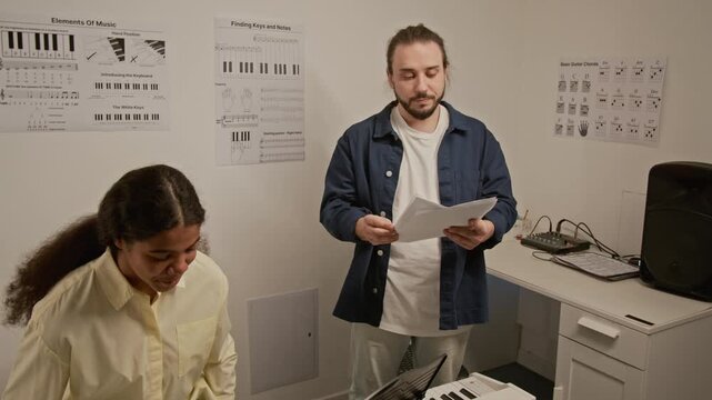 Zoom out shot of Caucasian male teacher greeting Black female student and placing sheet music in front of her after she sitting at digital piano. Clip for music school advertising