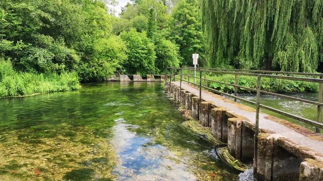 Fullerton Hampshire England UK. 29.06.2026. Video. A tranquil pathway crosses a clear river, showcasing vibrant foliage and the gentle movement of water over stones