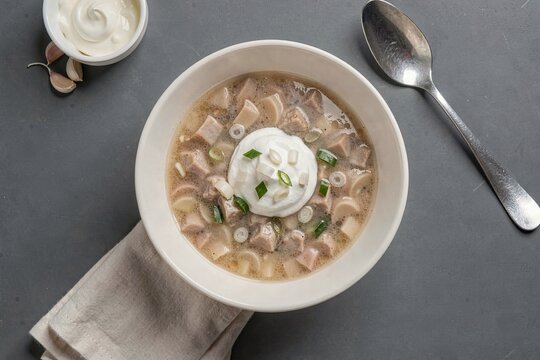 Bowl of Ciorba de Burta topped with garlic vinegar cream, garnished with green onions, on gray concrete surface. Concept of tripe soup with garlic vinegar cream served elegantly.