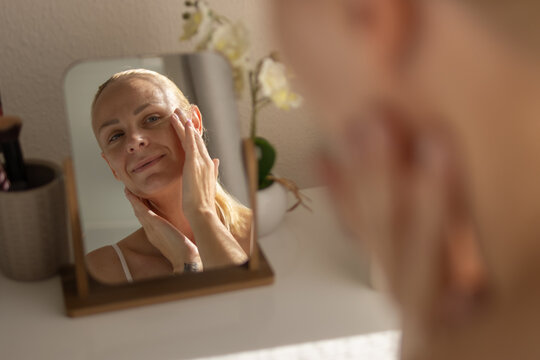 Woman smiling while applying face cream, moisturizing skin during daily self-care morning routine, reflecting freshness and wellness in mirror