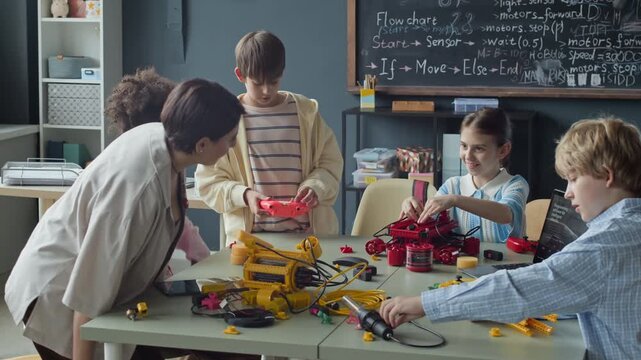 Multiethnic children building robots with laptop in classroom while female teacher joins them. Perfect for promoting STEM programs, school science classes, and educational technology