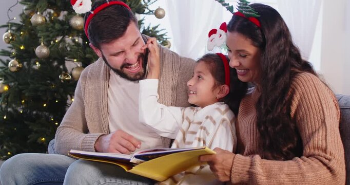Parents and child girl reading a holiday book. At home by festive Christmas tree, mother and father guide daughter as lights make warm atmosphere. Winter holidays celebration and family leisure. 