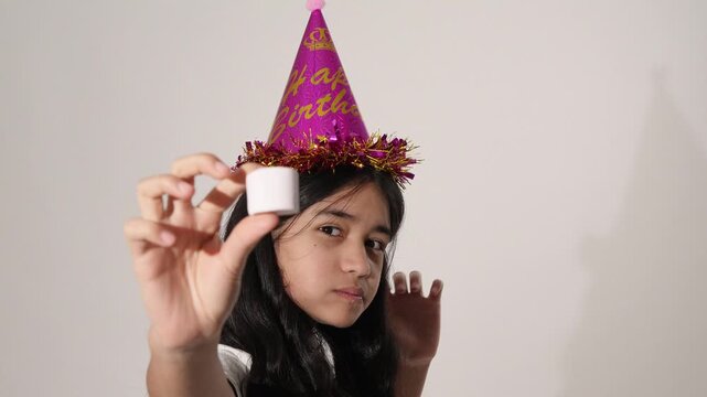 Young girl in a party hat eating a white marshmallow on white background