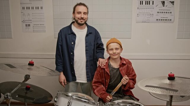 Zoom out portrait of young male teacher and teenager sitting at drum kit, smiling at camera. Ideal for promoting music schools and creative youth education