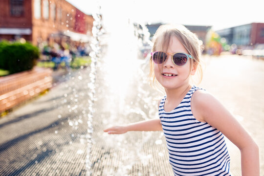 Cheerful little girl in sunglasses smiling next to a refreshing public water fountain stream on a hot sunny summer day