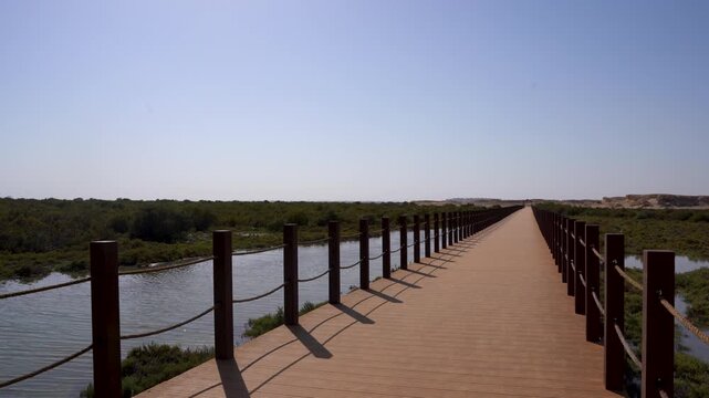 A wide landscape shot of the long wooden pedestrian boardwalk connecting the mainland to Purple Island (Jazirat Bin Ghannam) in Al Khor, Qatar. The pathway stretches through the natural wetland ecosys