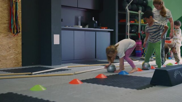 Young children participate in a fun fitness exercise, rolling balls between colorful cones in a gym. These playful children are developing their motor skills and coordination.