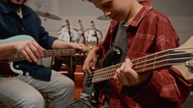 Male teenager learning to play bass guitar from male teacher in music studio. Footage for music school advertisements and youth education campaigns