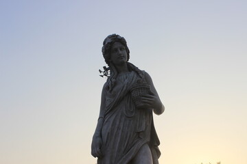 Fototapeta premium Stone statue of woman carrying harvest basket against soft pale twilight sky background.