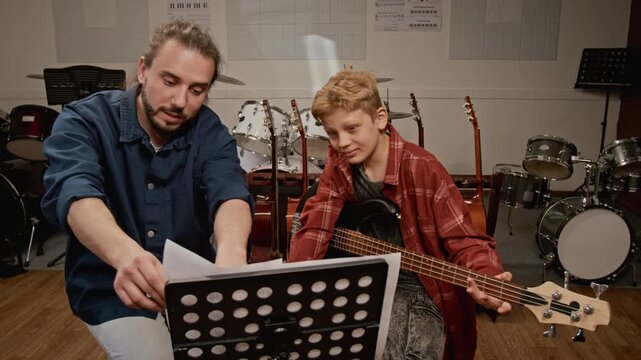 Young man teaching music to teenager boy holding bass guitar in studio. Footage for music school promos and private tutoring advertisements