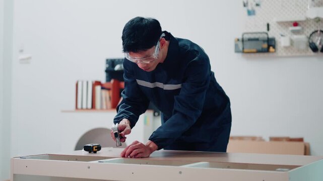 Asian Technician in Uniform Assembling Furniture with Tape Measure Indoors