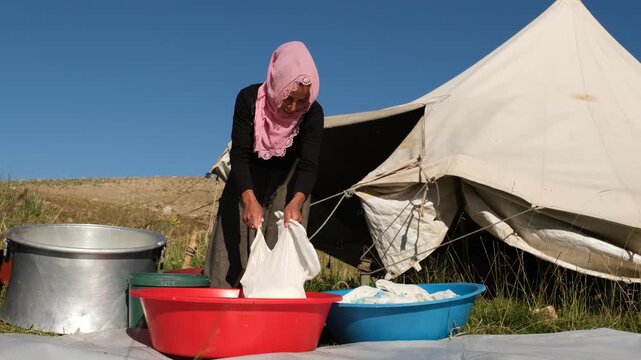 A woman lifts a long cheesecloth bag filled with curds above outdoor basins showing whey drainage, traditional cheese making, manual dairy work, and small-scale production in a sunny highland setting.