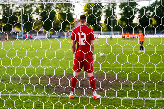 View through the net of a soccer goalkeeper in a red kit preparing to defend a penalty kick during a football match.