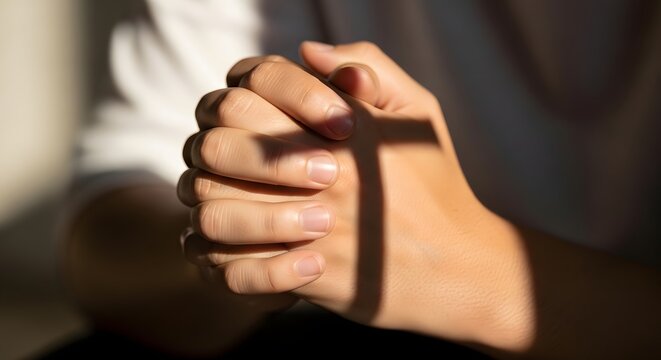 Close-up of human hands folded in prayer with a shadow of a christian cross cast over them as a symbol of faith and hope, religion