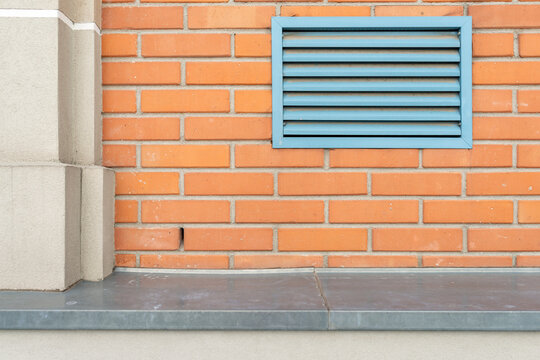 Front view of a red brick building facade featuring a blue rectangular ventilation grille and a zinc-covered ledge