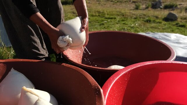 A woman squeezes a cloth bag of fresh curds over a red basin, showing a traditional cheese-making stage, manual whey draining, rural food preparation, and small-scale outdoor dairy production by hand.