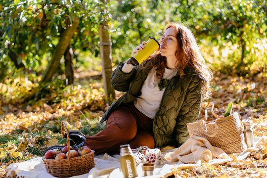 Relaxed red haired woman drinking water during sunny autumn picnic in apple orchard