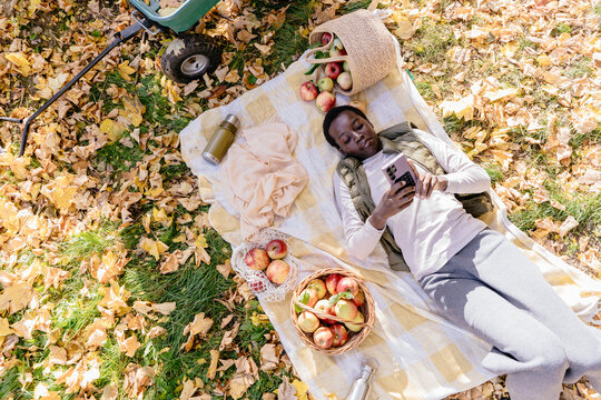 Relaxed Black woman using smartphone during autumn picnic after apple harvest
