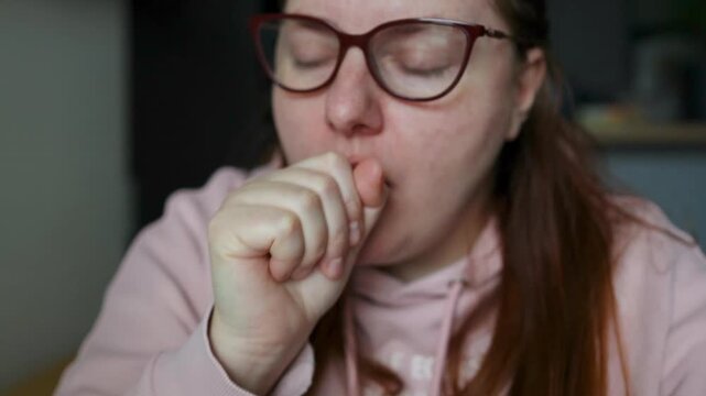A detailed portrait of a young Caucasian woman with red hair and glasses choking or coughing into her hand, showing physical discomfort and breathing difficulties during real-time video shot.