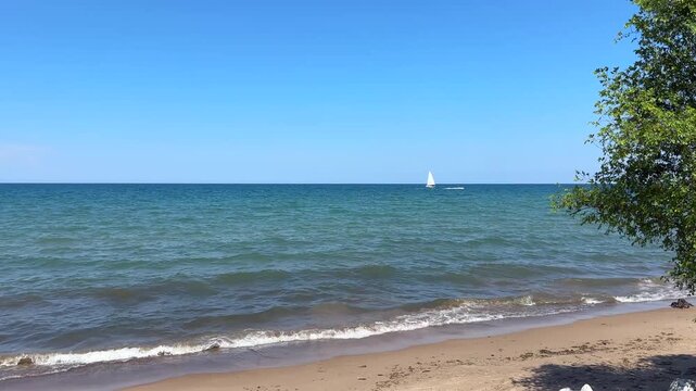 Waves in Lake Michigan washing up on a sandy beach with a sail boat in the distance on a sunny summer day