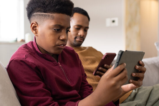 African American dad, teenage son sitting on couch at home, teen holding tablet, dad holding phone