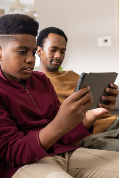 African father and adolescent son sitting on couch, holding smartphone and tablet, wearing pullover