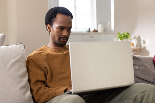 African man sitting on sofa at home using laptop, wearing brown sweater and neutral pants