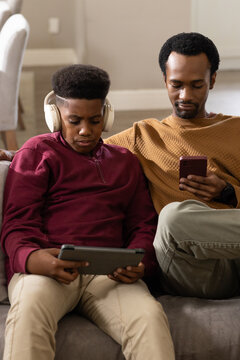 African American dad and teen son sitting on gray sofa holding tablet headphones and smartphone