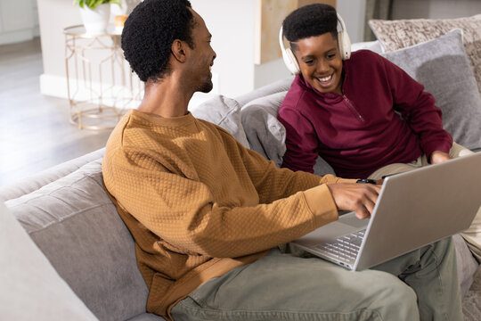 African American father pointing at laptop, teen son sitting on gray couch at home wearing headset