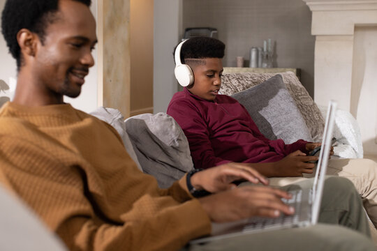 African American dad and teen son sharing sofa at home using laptop and device with headphones