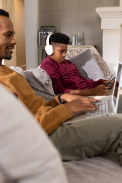 Father and teenage son on living room couch, father typing laptop, teen in burgundy using phone
