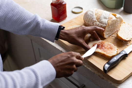 African American man wearing knit sweater and wristwatch spreading jam on bread at kitchen counter