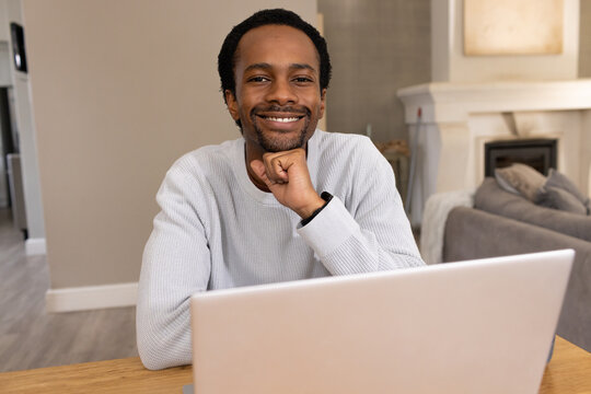African man sitting at wooden table in living room using laptop and smiling with watch