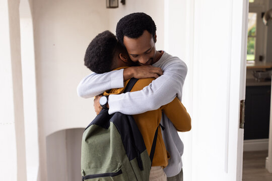 African father hugging teenage son in home entryway, showing olive-green backpack and wristwatch