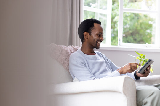 Adult African American man wearing light blue sweater sitting at home tapping tablet with watch