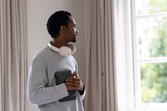 Man standing on left by multi-pane window looking out holding tablet and wearing headphones