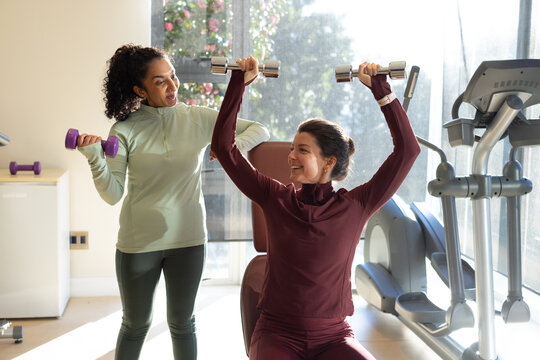 Diverse female trainer and client working out in fitness room with chrome dumbbells on padded bench