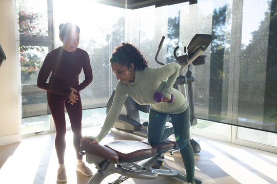 Female coach spotting African-American client in sportswear doing purple dumbbell row on home bench