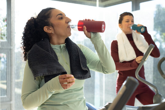 Diverse female gym partners wearing workout gear drinking from reusable bottles on cardio machines