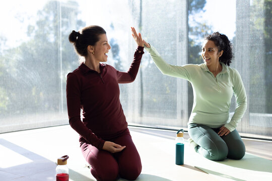 Female friends kneeling on yoga mats in sportswear at studio giving high-five near water bottles
