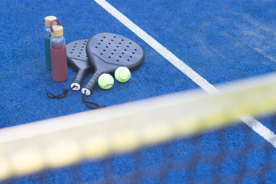Padel rackets are lying crossed on blue court with blurred net, balls and three water bottles