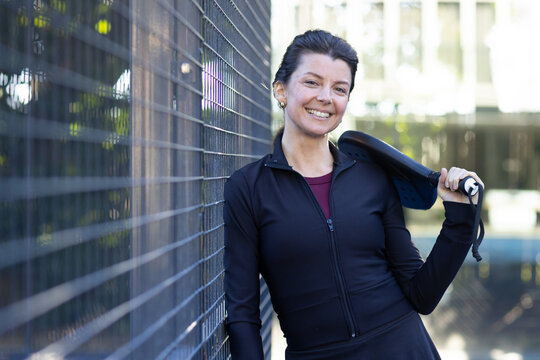 Adult female wearing black jacket leaning against chain-link fence at court holding paddle, smiling
