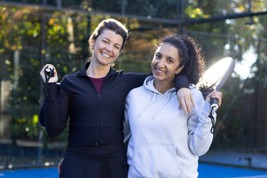 Diverse women friends standing arm-in-arm on blue court holding paddles with straps in sportswear