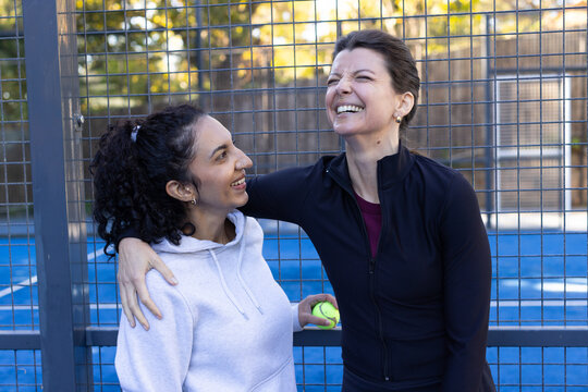 Female friends laughing and hugging on blue fenced sports court wearing hoodies holding tennis ball