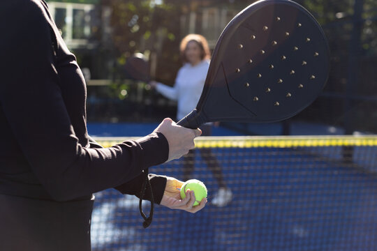 Diverse female duo serving on blue court in black-top holding black-paddle, yellow-ball, copy space