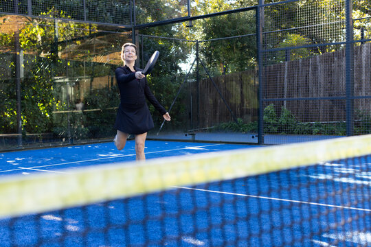 Woman swinging padel racket and lunging on blue padel court past yellow net, wearing black top