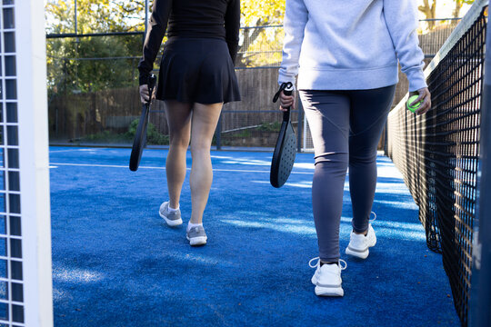 Diverse female friends walking on blue paddle court in sportswear carrying paddles and green ball