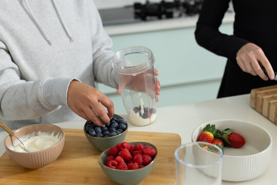 Two female friends preparing blueberry yogurt smoothie at kitchen counter with clear blender cup