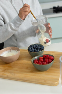 Yogurt and berries are being stirred in clear mixing cup using wooden spoon on kitchen countertop